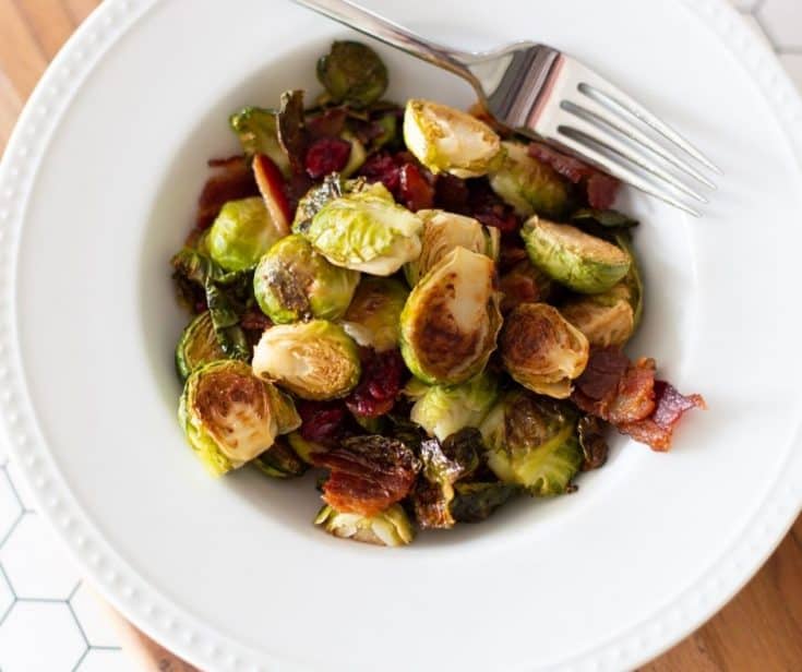 roasted Brussel sprouts with bacon and cranberries in a white bowl with fork on the side by The Moments At Home