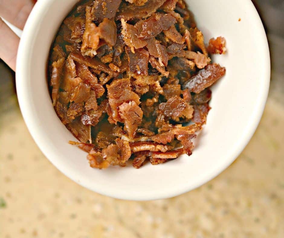 bowl of crispy bacon pieces over a skillet with creamy garlic sauce simmering