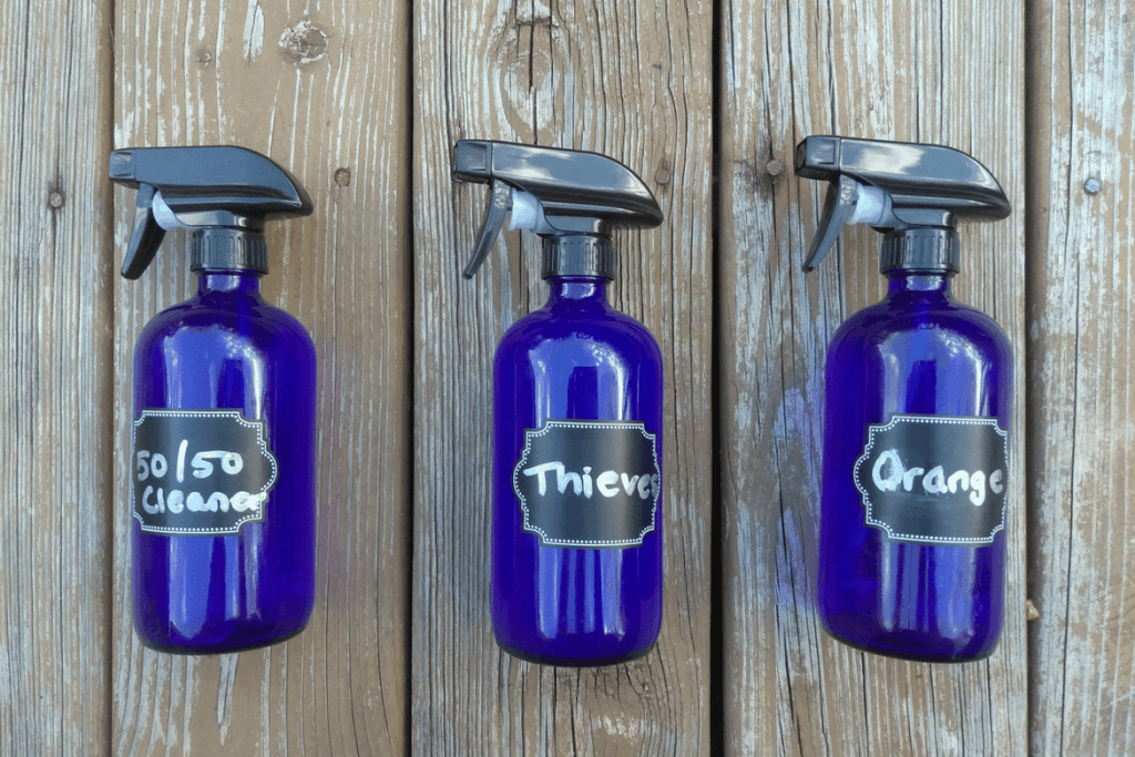 3 blue glass bottles holding homemade non-toxic cleaning solutions with spray tops and labels saying "50/50, thieves, and orange"